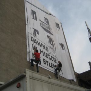 Wyklejanie szablonu muralu na elewacji budynku przy wykorzystaniu technik alpinistycznych.