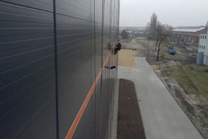 Malowanie pasów szablonowych loga na zewnętrznej ścianie hali produkcyjnej przy wykorzystaniu technik alpinistycznych.