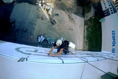 Wyklejanie szablonu loga na zewnętrznej elewacji hali produkcyjnej przy wykorzystaniu technik alpinistycznych.