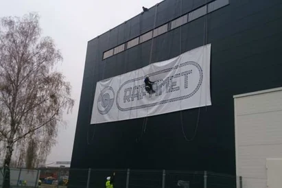 Wyklejanie szablonu loga na zewnętrznej ścianie hali produkcyjnej przy wykorzystaniu technik alpinistycznych.