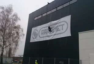 Wyklejanie szablonu loga na zewnętrznej ścianie hali produkcyjnej przy wykorzystaniu technik alpinistycznych.